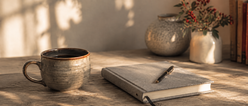 A ceramic mug of coffee on a desk with a notebook and pen, all in muted colors.